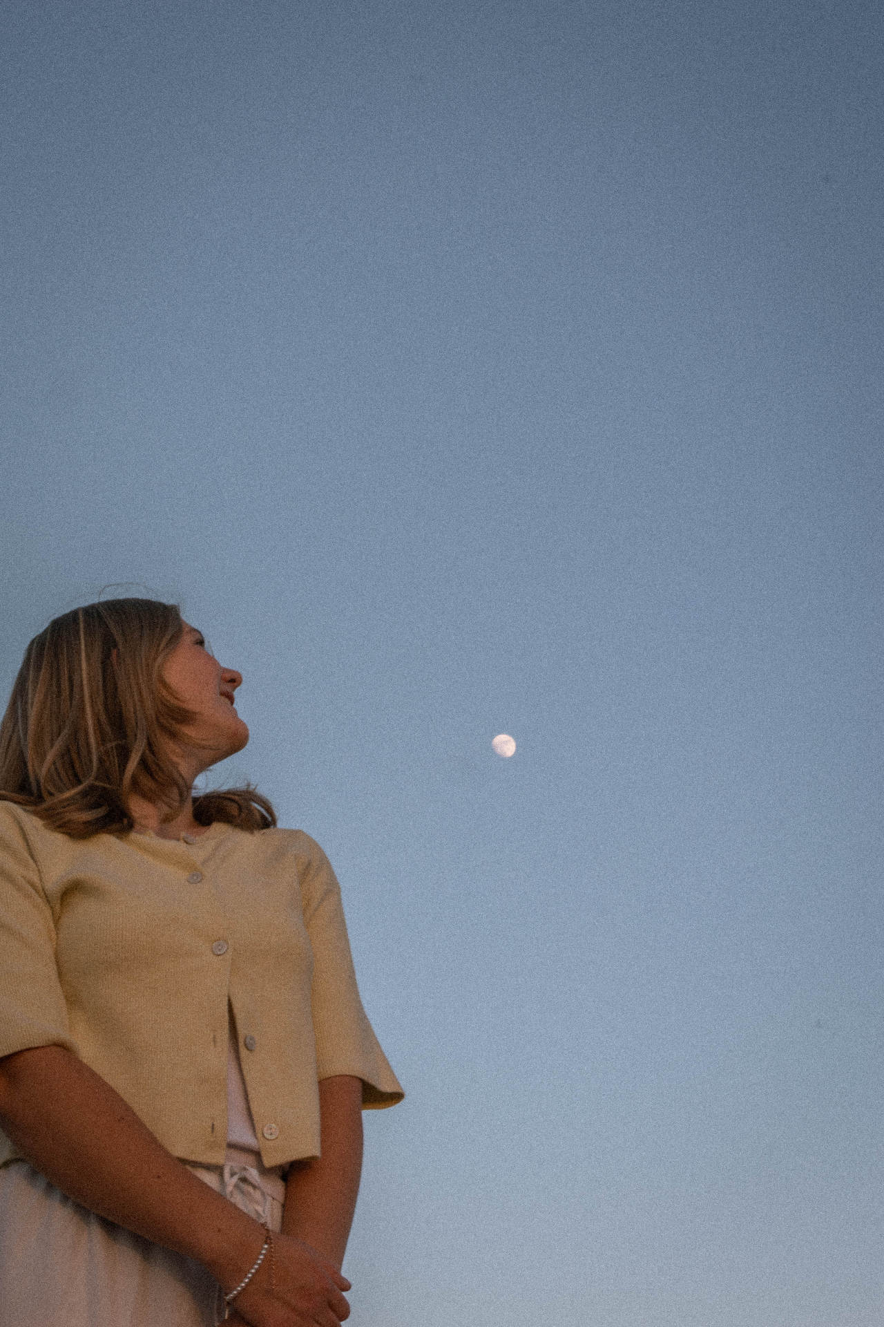 Girl in a yellow cardigan looking up at a daytime moon in a soft blue sky