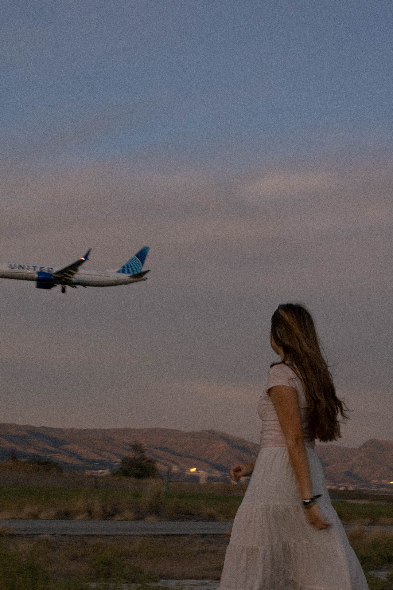 Girl in a white dress watching a United airplane fly over the foothills at dusk