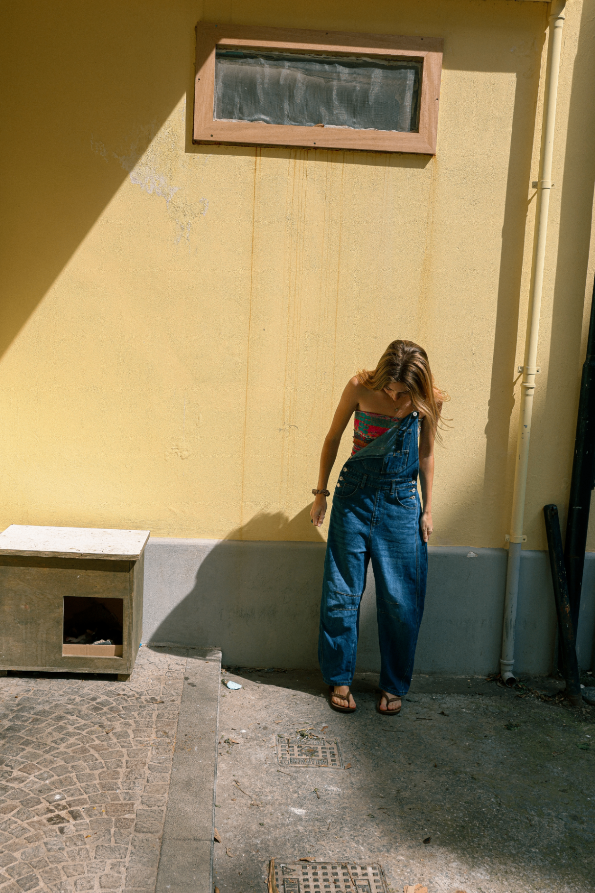 Young woman in denim overalls leaning against a sunny yellow stucco wall, hair falling over her face